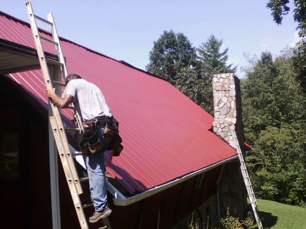 Roofer on a ladder beside a red metal roof and stone chimney — Soffit & Fascia Repair by Roofing Brothers in Ephraim